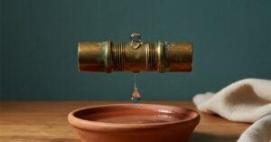 A short length of aged brass pipe with a threaded joint dripping water into a shallow terra cotta dish on an oak workbench against a deep teal wall