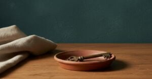 A single aged brass key resting in a shallow terra cotta dish on an oak workbench against a deep teal plaster wall